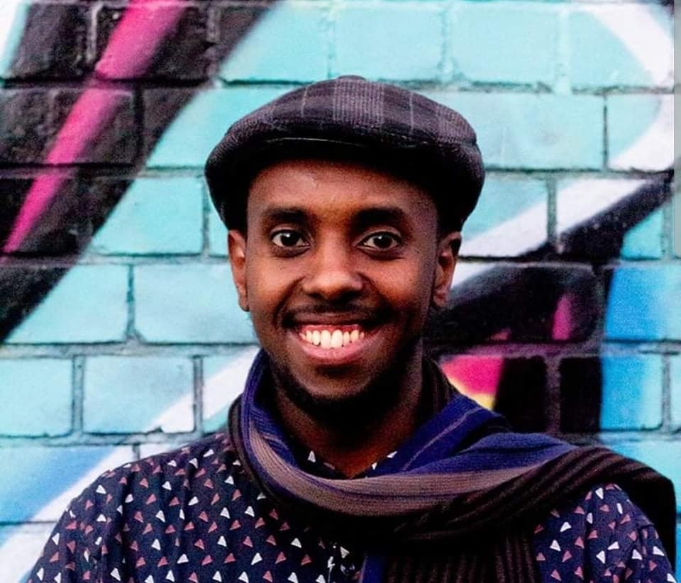 Writer, Awale Ahmed, standing front on wearing a blue top with a pattern on small white and purple triangles, a scarf and hat. They are smiling and standing in front of a graffiti wall.