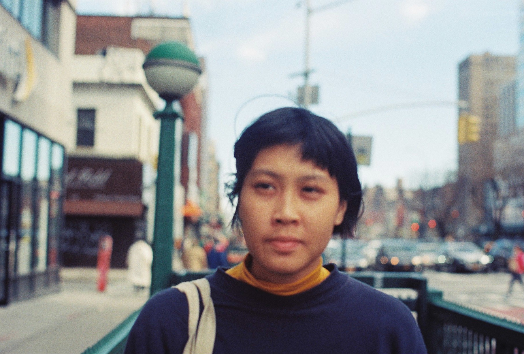 A headshot of Thao Ly, taken on the street. Thao is wearing a dark blue sweater and has a light coloured tote over her right shoulder. Thao looks off camera to the left. The street is blurred behind her.