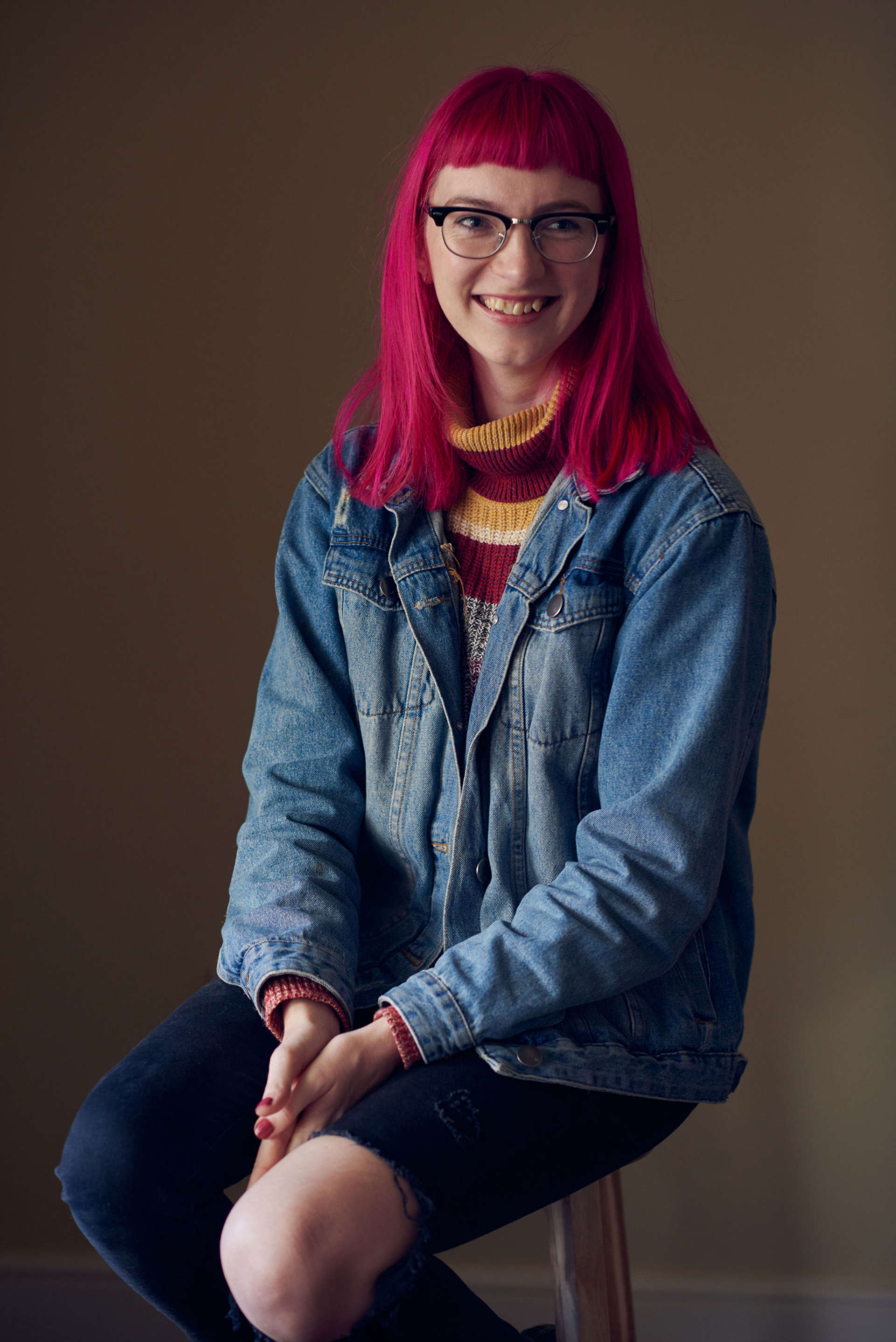A headshot of the writer Josephine Newman. Jospehine has long dark pink hair and is wearing glasses. She is smiling at the camera.