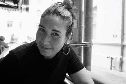 A black and white portrait of the writer Anne-Marie Te Whiu. Anne-Marie wears a dark short-sleeved tshirt and her hair is pulled up. She wears hoop earrings and smiles at the camera.