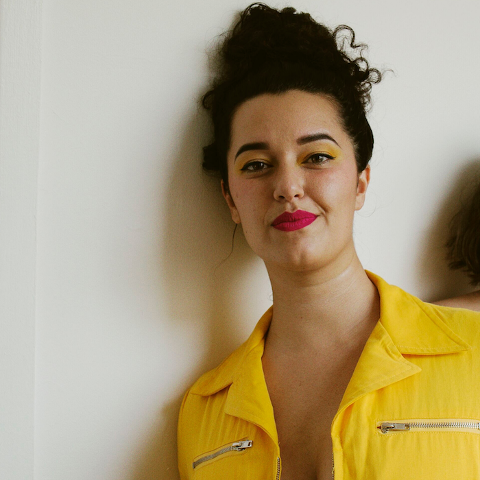 A headshot of Jessican Paraha wearing a yellow collared shirt, red lipstick, yellow eyeshadow, with her dark curly hair in a bun on top of her head. She is leaning her right shoulder against a white wall looking at the camera and smiling slightly.