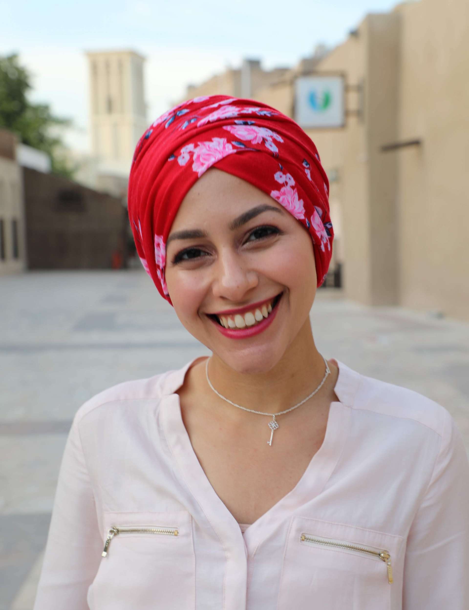 A headshot of Lur Alghurabi wearing a white long sleeved shirt, silver necklace, red lipstick and a red scarf with pink flowers wrapped around her head. She is smiling at the camera and the background is of a blurry outside setting.