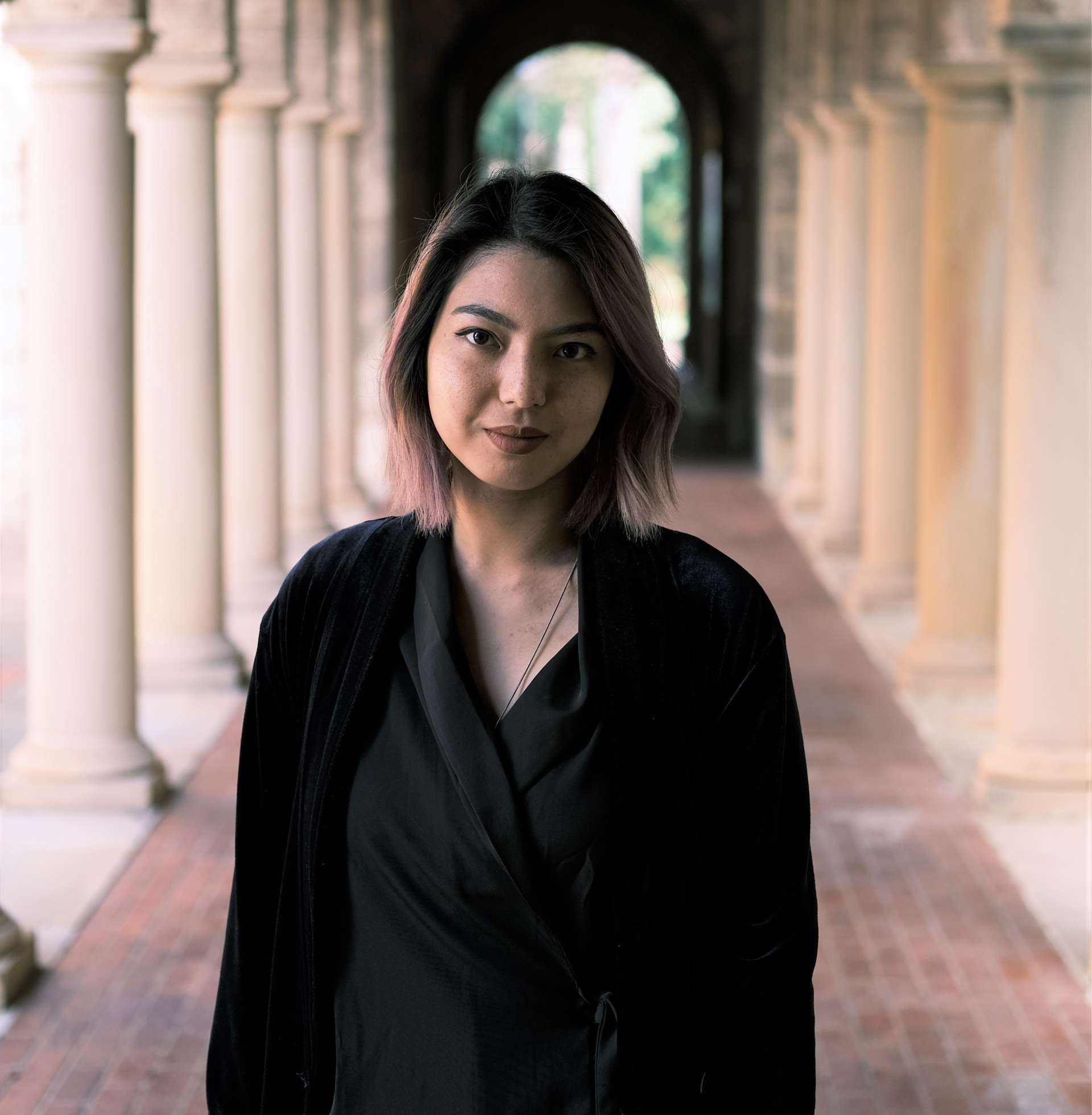 A headshot of the writer Prema Arasu. Prema is standing in the middle of a long outdoor hallway, framed by lines of stone columns. She has long dark shoulder-length hair that has hints of blonde and pink at the ends. Prema is wearing a black top and black cardigan. She looks to the camera with a closed-mouth smile.